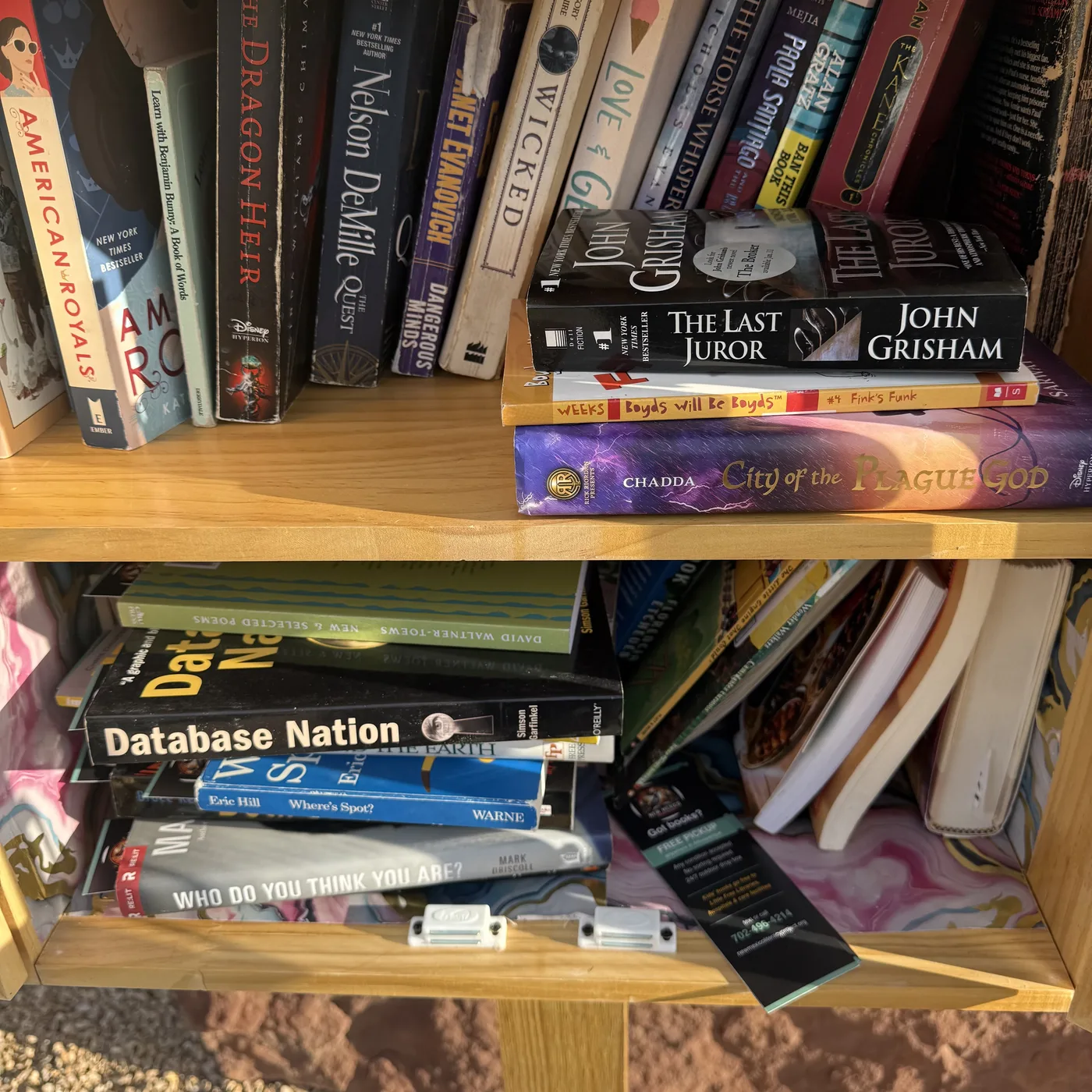 An indoor wooden bookshelf restocked with NMLP-donated books. Top shelf visible: American Royals YA, Learn with Benjamin Bunny, The Dragon Heir, Nelson DeMille's The Quest, Janet Evanovich, Wicked, Love & Gelato, John Grisham's The Last Juror stacked, Boyds Will Be Boyds, Chadda's City of the Plague God (Rick Riordan Presents), Janet Evanovich, Paola Santiago, Alan Gratz Ban This Book, The Kane Chronicles, more. Bottom shelf: Database Nation by Simson Garfinkel (O'Reilly), Where's Spot? by Eric Hill (Warne), Mark Driscoll Who Do You Think You Are? Bottom right: NMLP black bookmark visible showing 'Got books? Free pickup' and the 702-496-4214 phone number.