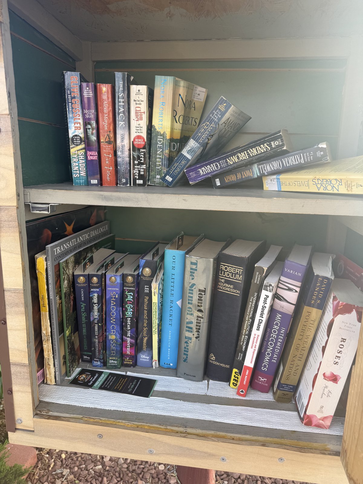 Two-shelf Little Free Library filled with donated books. Top shelf has adult commercial fiction — Clive Cussler Shadow Tyrants, Nora Roberts Identity, Tom Clancy Every Man a Tiger, The Shack, Amanda Quick, Sandra Brown Charade, Patricia Cornwell Point of Origin. Bottom shelf mixes more Rick Riordan Presents middle-grade titles including Aru Shah and the Tree of Wishes, The Shadow Crosser, and Pahua and the Soul Stealer with academic titles including Transatlantic Dialogue, Intermediate Microeconomics, and The Wealth and Poverty of Nations. A New Mexico Literacy Project bookmark rests on the lower shelf.