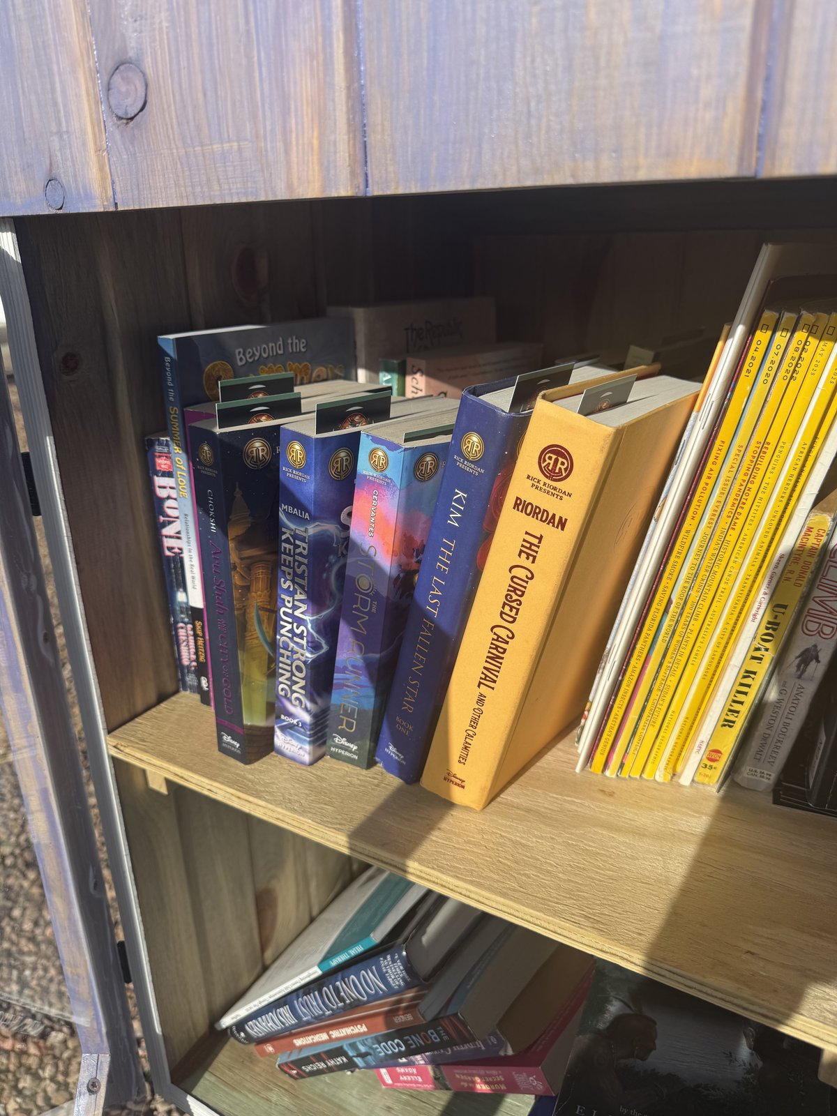 Cedar-frame Little Free Library stocked with Rick Riordan Presents middle-grade hardcovers — Tristan Strong Keeps Punching, The Storm Runner, Aru Shah, Sal and Gabi, The Last Fallen Star, The Cursed Carnival — next to a shelf of vintage National Geographic magazines. New Mexico Literacy Project bookmarks are visible tucked into the hardcover spines. Albuquerque North Valley, April 2026.