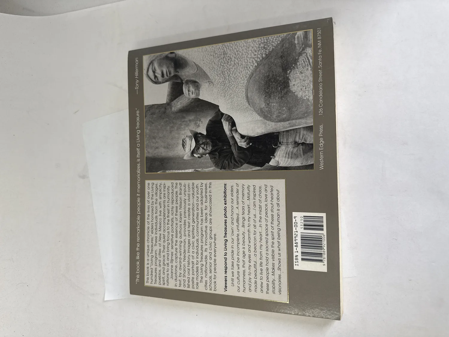 Back cover of Living Treasures showing Tony Hillerman blurb calling the book itself a Living Treasure, duotone portrait photos of New Mexican elders, description of the Living Treasures program honoring over one hundred elders from villages pueblos and cities of New Mexico, Western Edge Press 126 Candelario Street Santa Fe NM 87501, ISBN barcode. Photographed at the New Mexico Literacy Project sorting desk in Albuquerque.