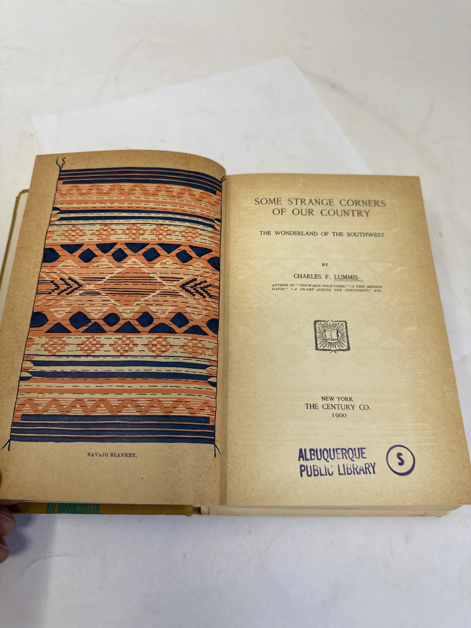 Title page spread of Some Strange Corners of Our Country by Charles F. Lummis, The Century Co. 1900, with Navajo blanket color plate frontispiece and Albuquerque Public Library stamp visible. Photographed at the New Mexico Literacy Project sorting desk in Albuquerque.