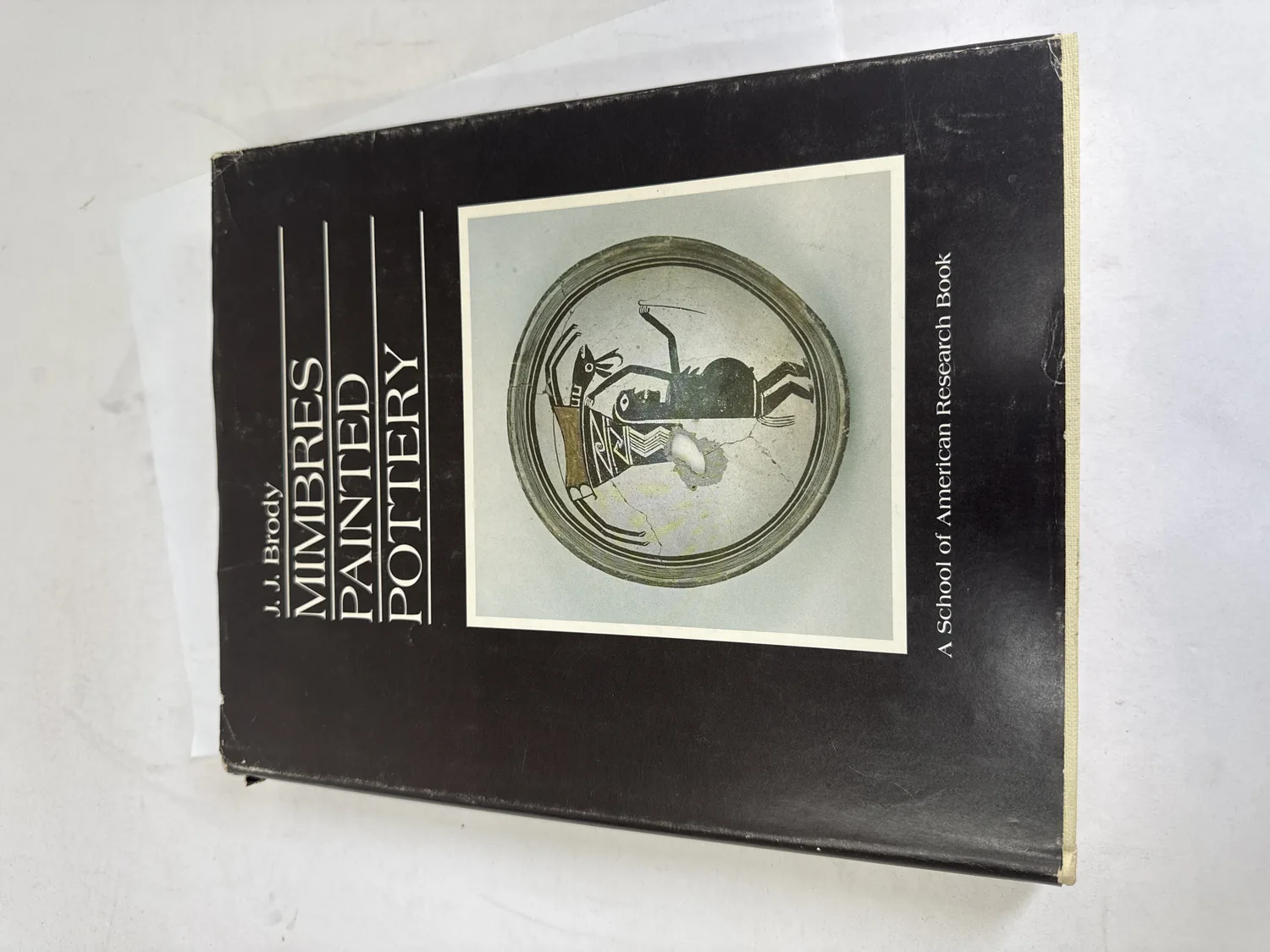 Front cover of Mimbres Painted Pottery by J.J. Brody, first edition 1977, showing a classic Mimbres black-on-white figurative bowl with an insect or grasshopper figure, published as A School of American Research Book by University of New Mexico Press. Photographed at the New Mexico Literacy Project sorting desk in Albuquerque.