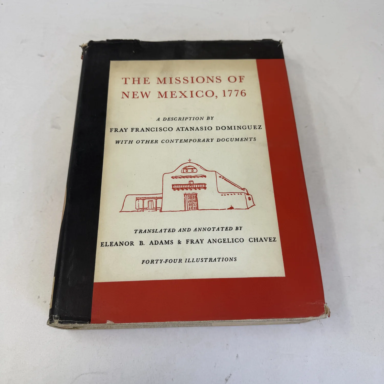 Front of the dust jacket on the 1956 first edition of The Missions of New Mexico, 1776, red, black and cream with a line drawing of a mission church and the words Forty-four Illustrations, photographed at New Mexico Literacy Project