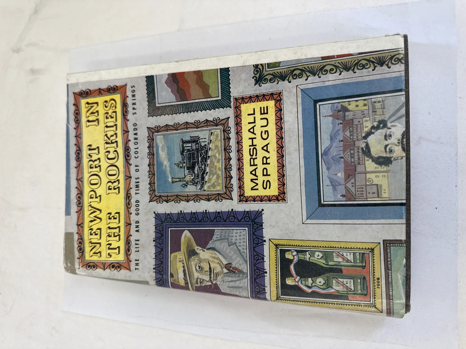 Front cover and dust jacket of Newport in the Rockies by Marshall Sprague, Sage Books and Alan Swallow 1961. Decorative stamp-style illustrations of Colorado Springs scenes including Pikes Peak, the Broadmoor, horse-drawn carriages, and mining camps on a cream background. Photographed at the New Mexico Literacy Project sorting desk in Albuquerque.