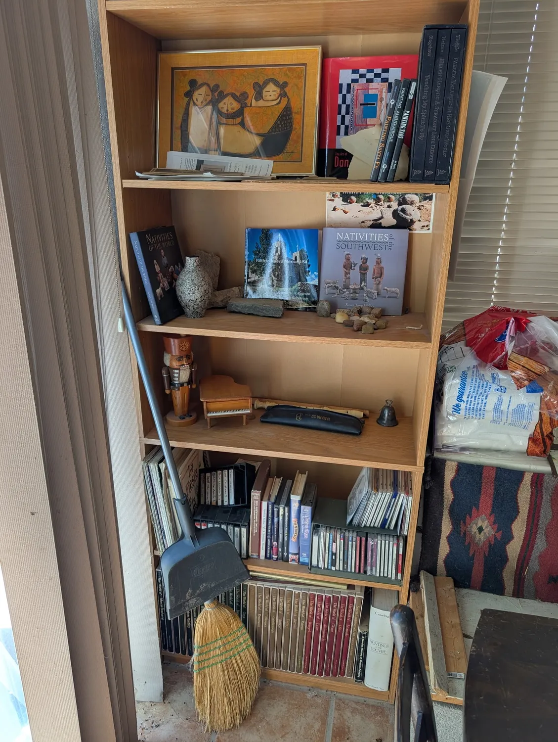 A porch bookshelf in a New Mexico home with a Pueblo-style painting of seated figures on the top shelf, Nativities of the Southwest book, a hand-carved kachina figure, pottery, cassette tapes, and books on the lower shelves — photographed during a book and media cleanout by Josh Eldred of the New Mexico Literacy Project in Albuquerque, with a Southwestern rug visible to the right