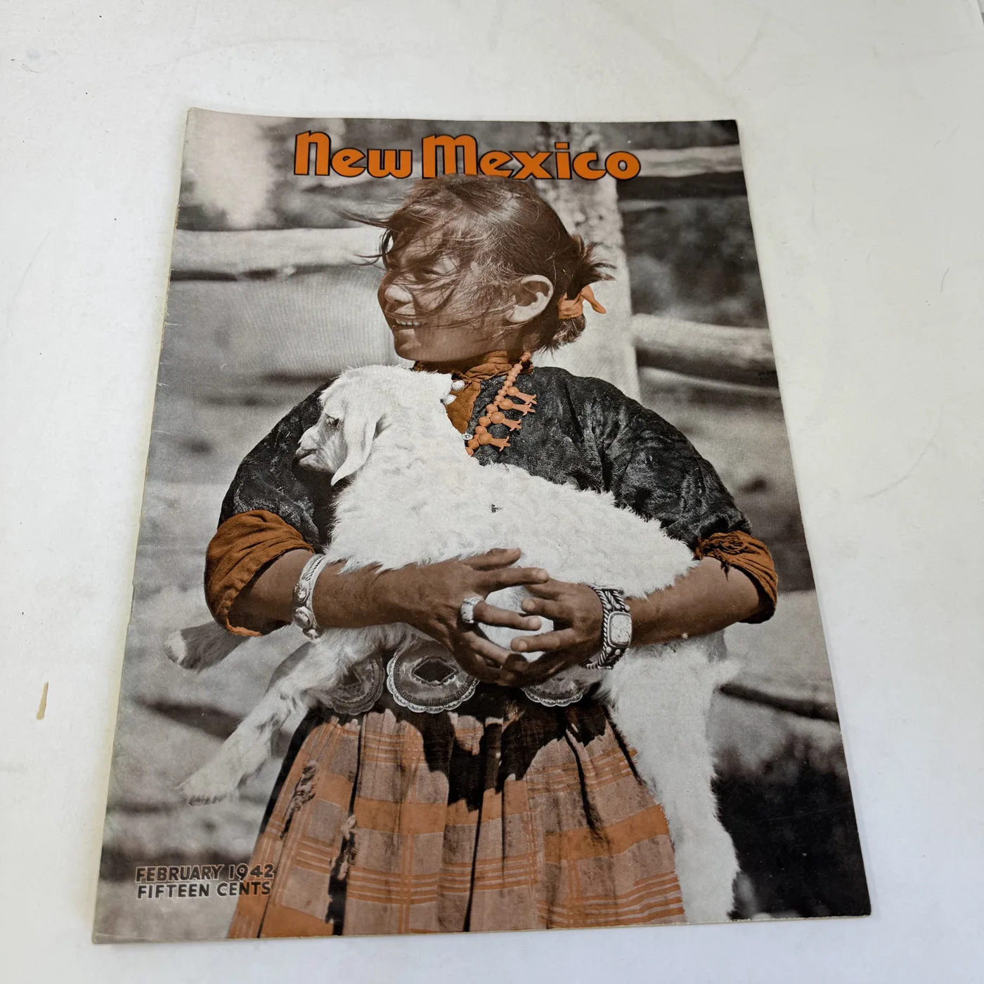 New Mexico Magazine February 1942 cover: hand-tinted photograph of a Navajo girl carrying a white lamb, wearing stamped-silver bracelets and a concho belt — photographed at New Mexico Literacy Project