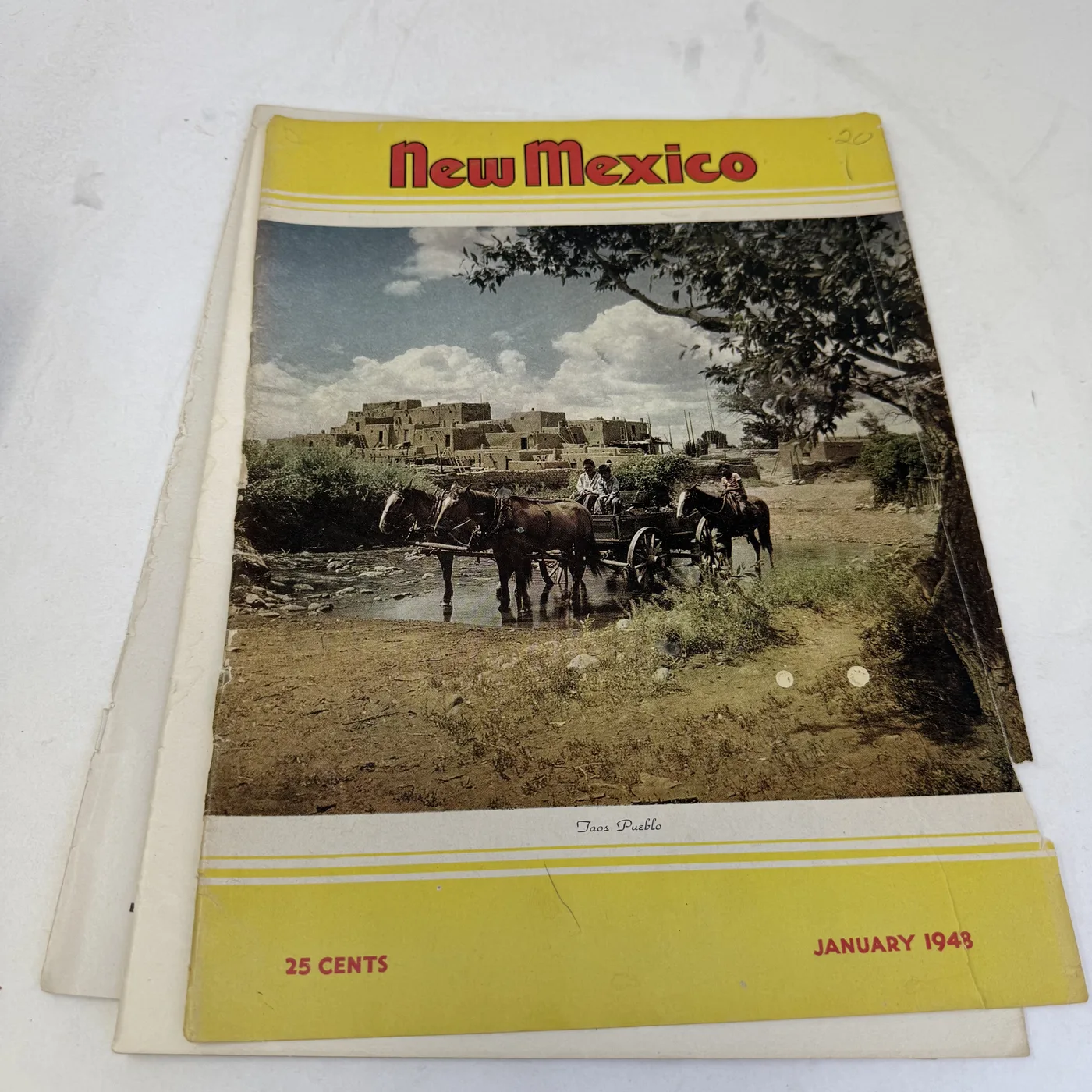 New Mexico Magazine January 1949 cover: horse-drawn wagon fording a shallow river, issue fanned on top of other issues from the run — photographed at New Mexico Literacy Project