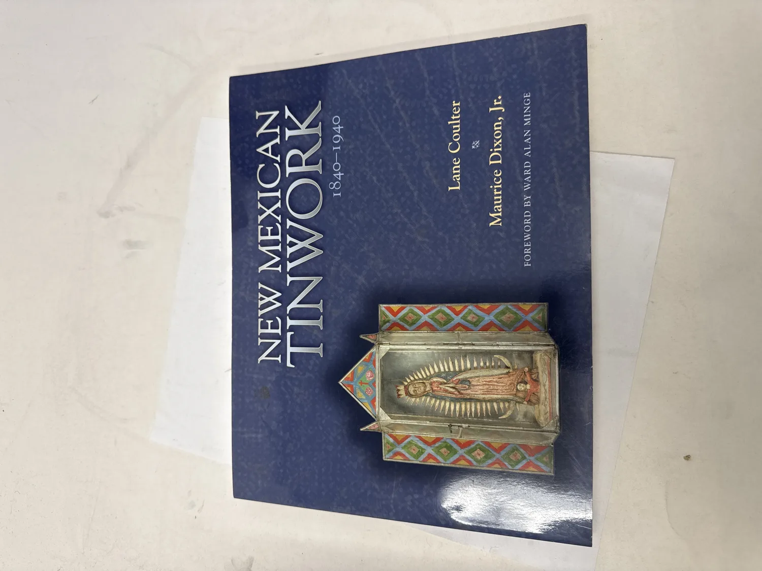 Front cover of New Mexican Tinwork 1840-1940 by Lane Coulter and Maurice Dixon Jr., navy blue field with silver typography and a full-color photograph of a tin nicho shrine box containing a painted santo figure in warm earth tones, University of New Mexico Press 1990 first edition. Photographed at the New Mexico Literacy Project sorting desk in Albuquerque.