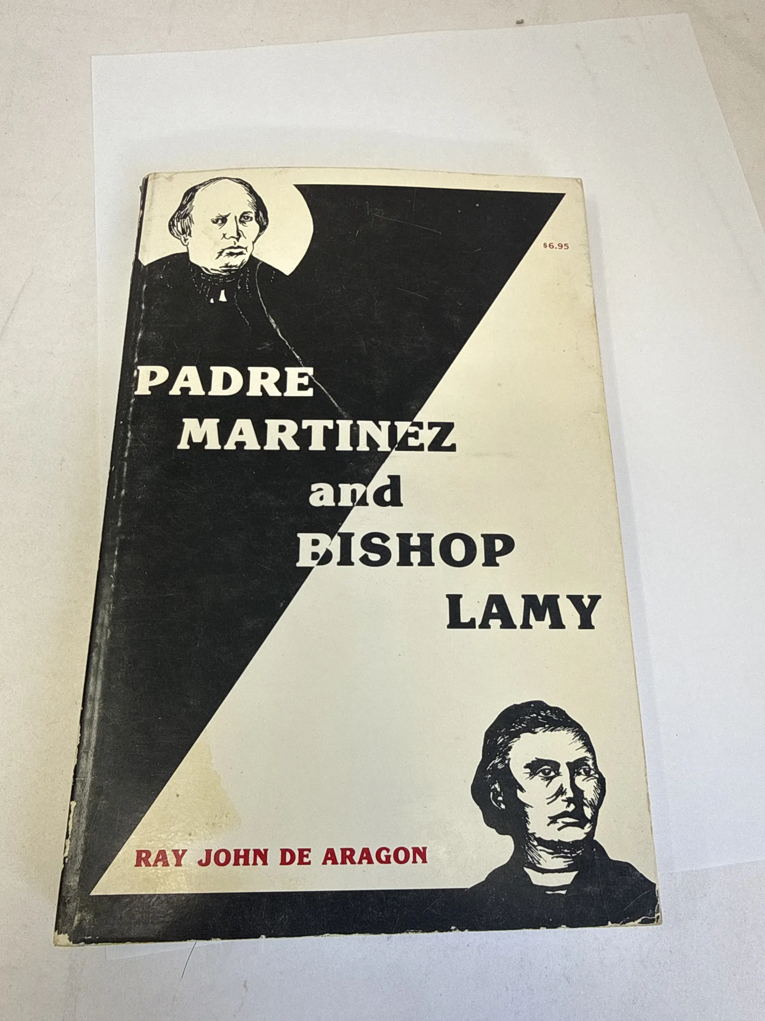 Cover of Padre Martinez and Bishop Lamy by Ray John de Aragón showing bold black-and-white graphic design with portraits of both Padre Martínez and Bishop Lamy, $6.95 price visible on the spine, published by Pan-American Publishing Company Las Vegas New Mexico 1978. Photographed at the New Mexico Literacy Project sorting desk in Albuquerque.