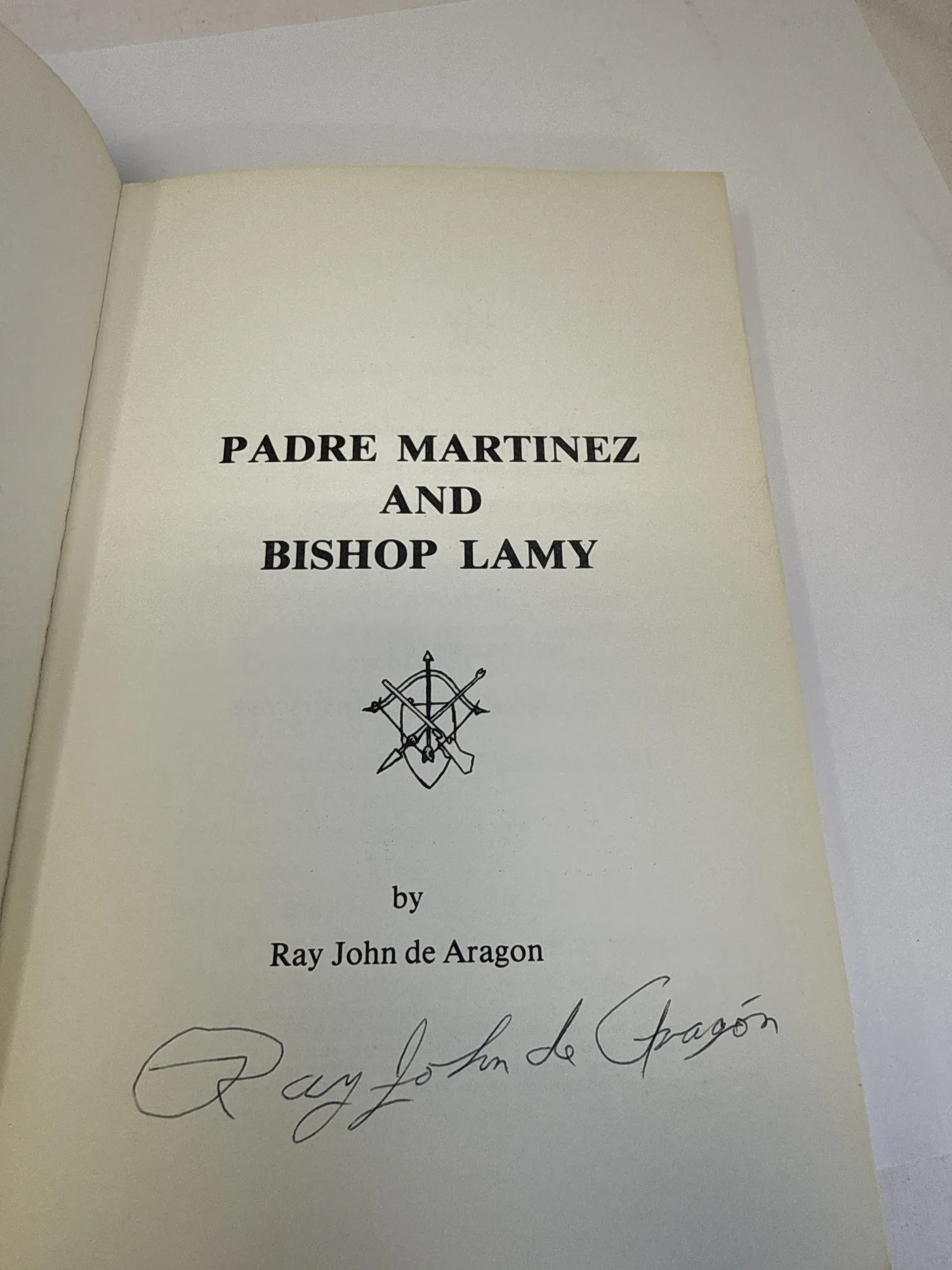 Title page of Padre Martinez and Bishop Lamy signed by Ray John de Aragón in black ink, with crossed-keys religious symbol between the title and author byline, Pan-American Publishing Company Las Vegas New Mexico 1978. Photographed at the New Mexico Literacy Project sorting desk in Albuquerque.