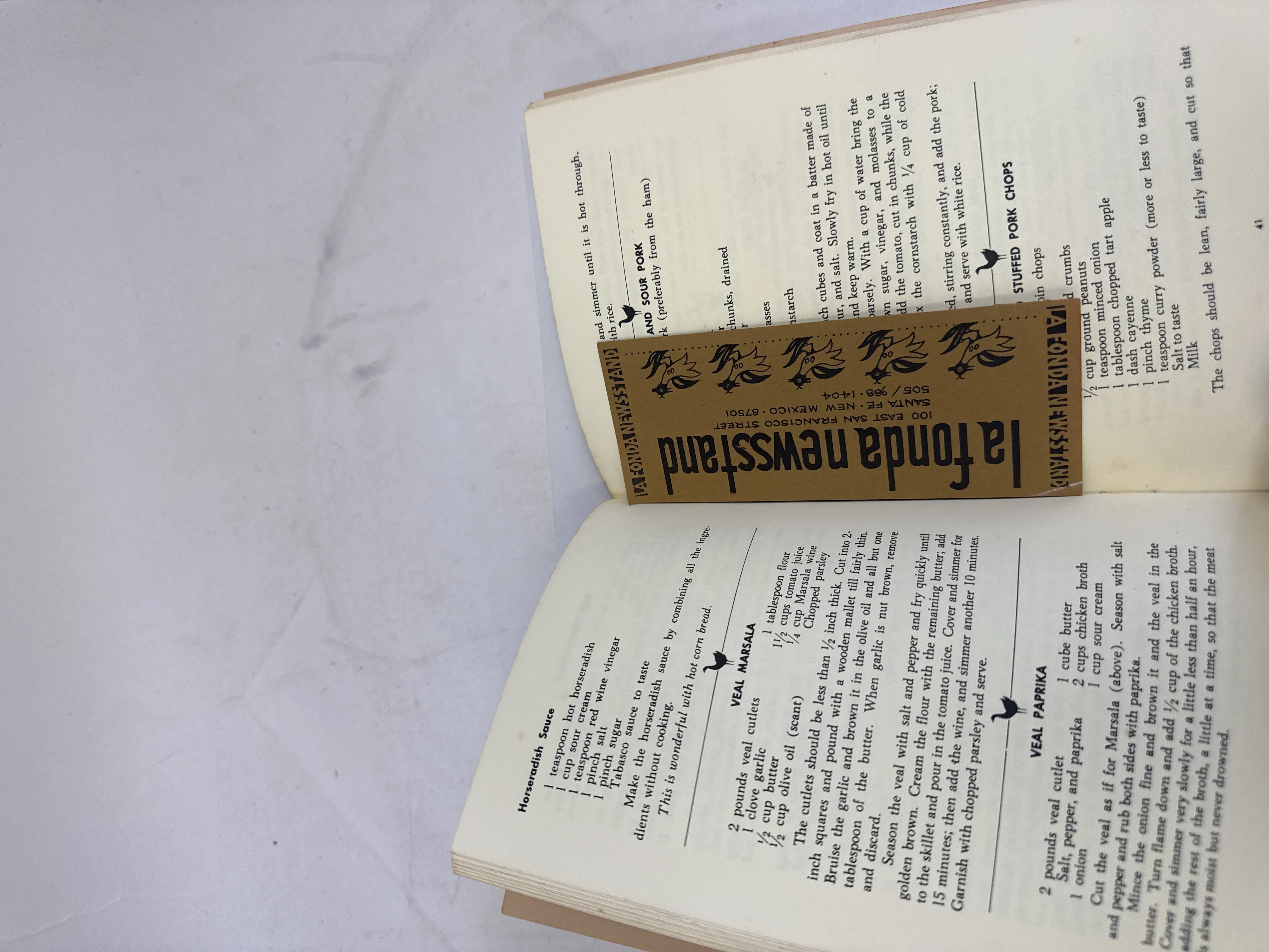 Interior recipe spread from the Meat section of Cooking with a Silver Spoon showing Veal Marsala and Veal Paprika recipes, with a La Fonda Newsstand bookmark tucked in as a placeholder. The bookmark shows la fonda newsstand in decorative type with bird motifs, 100 East San Francisco Street, Santa Fe, New Mexico 87501, phone 505-988-1404. Provenance of purchase at La Fonda Hotel on the Santa Fe plaza, circa early 1970s.