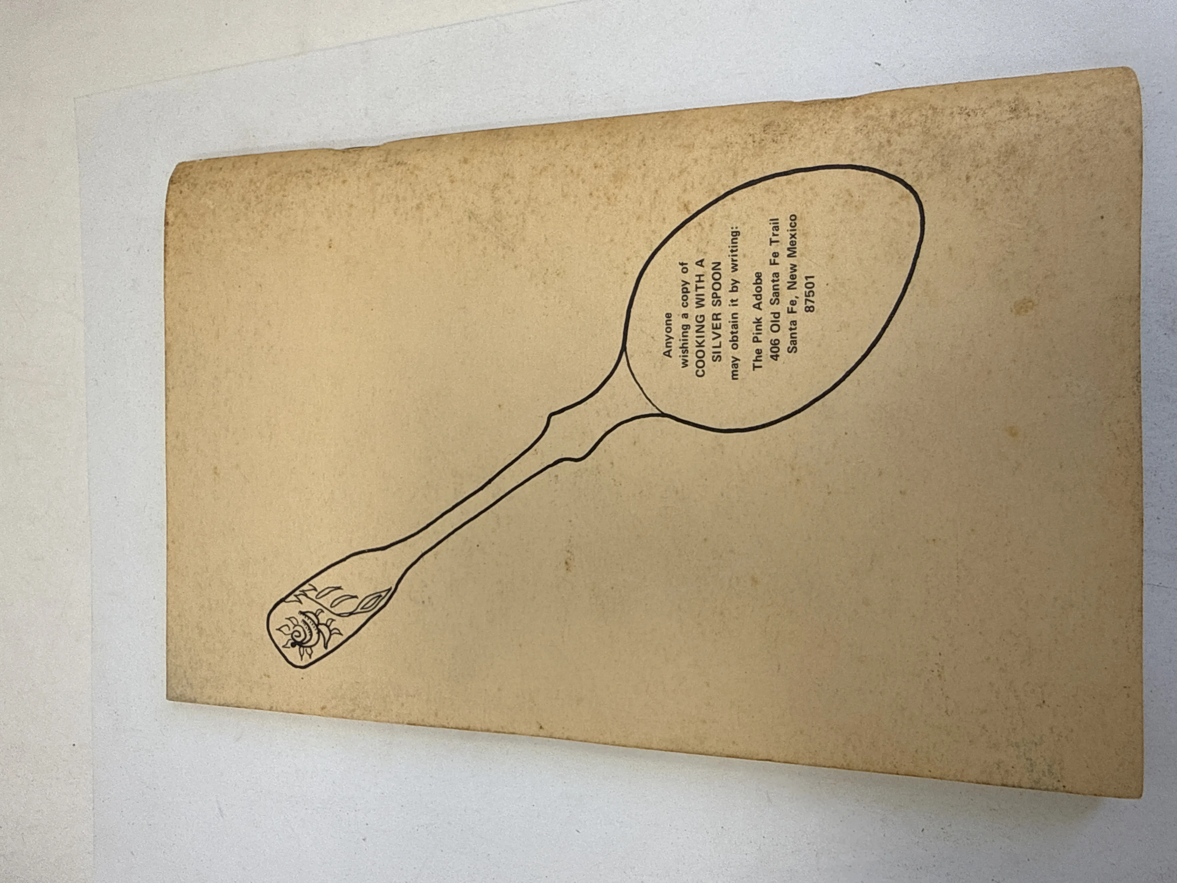 Back cover of Cooking with a Silver Spoon by Rosalea showing a line drawing of an ornate silver spoon with rose pattern on handle. Text inside the spoon bowl reads: Anyone wishing a copy of Cooking with a Silver Spoon may obtain it by writing The Pink Adobe 406 Old Santa Fe Trail Santa Fe New Mexico 87501. Age spotting on cream paper.