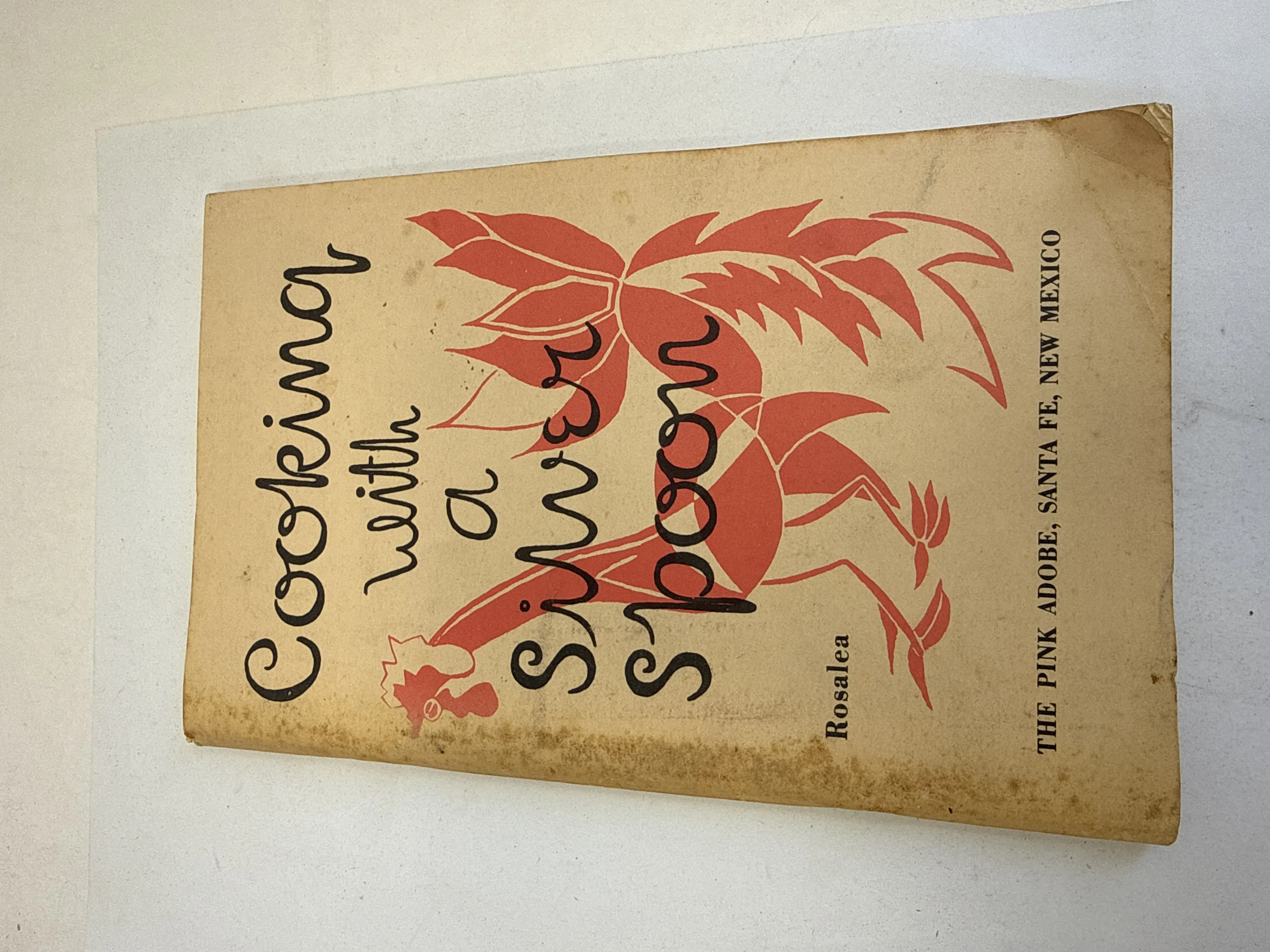 Front cover of Cooking with a Silver Spoon by Rosalea, self-published by The Pink Adobe restaurant, Santa Fe, New Mexico, 1970. Cream tan softcover with age toning, red stylized bird and plant illustration, handwritten-style title lettering. Rosalea and The Pink Adobe Santa Fe New Mexico printed along the right edge. Found in a New Mexico estate cleanout by the New Mexico Literacy Project.