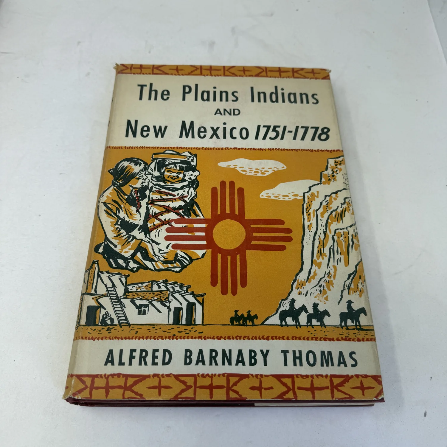Pictorial dust jacket of the 1940 first edition of The Plains Indians and New Mexico, 1751-1778 by Alfred Barnaby Thomas, yellow-orange with a red Zia sun symbol, a pueblo and riders on horseback, photographed at New Mexico Literacy Project