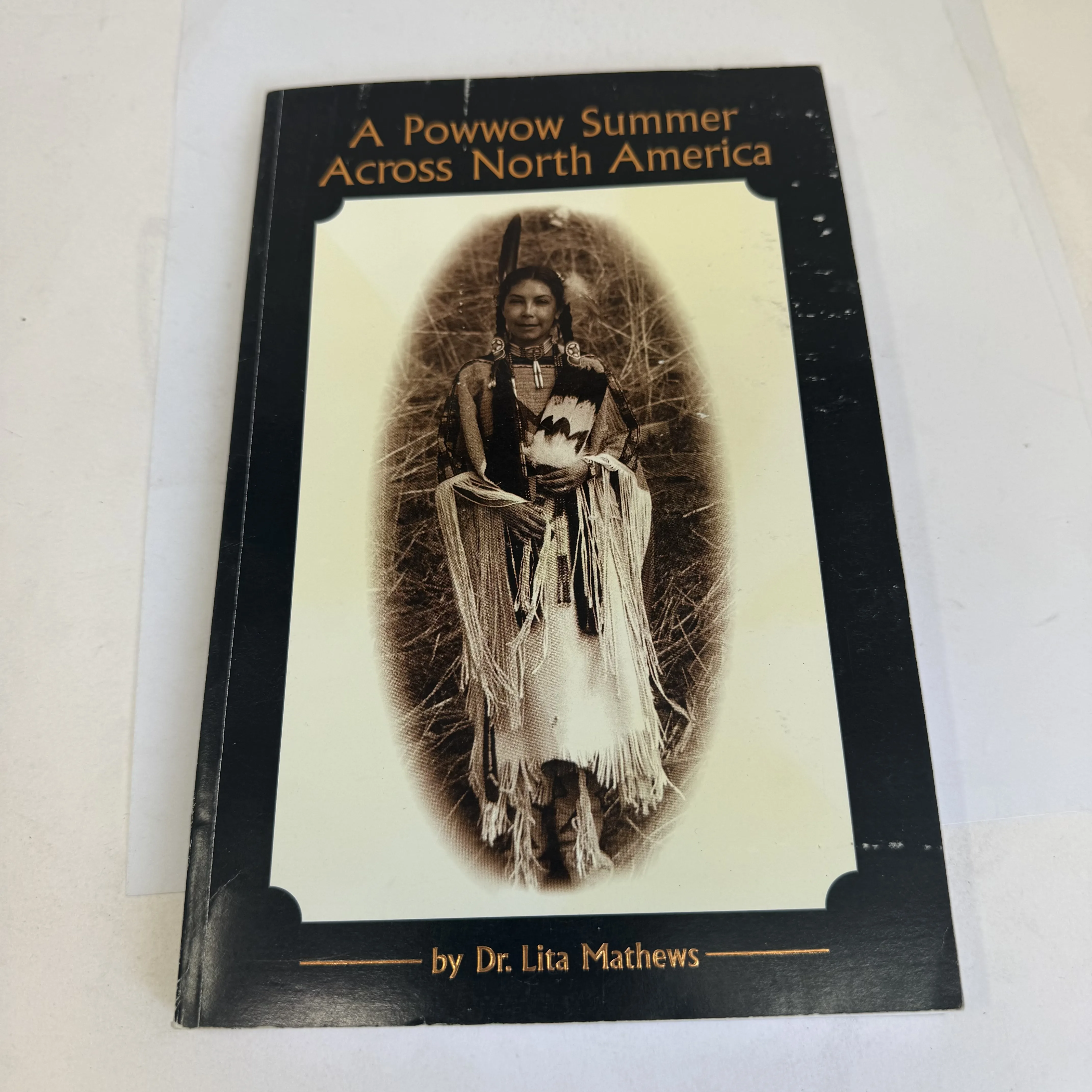 Front cover of A Powwow Summer Across North America by Dr. Lita Mathews — black cover with sepia-toned oval photograph of a Native American dancer in traditional buckskin regalia with fringe, feather, and beadwork, gold lettering, A Gathering of Nations Publication