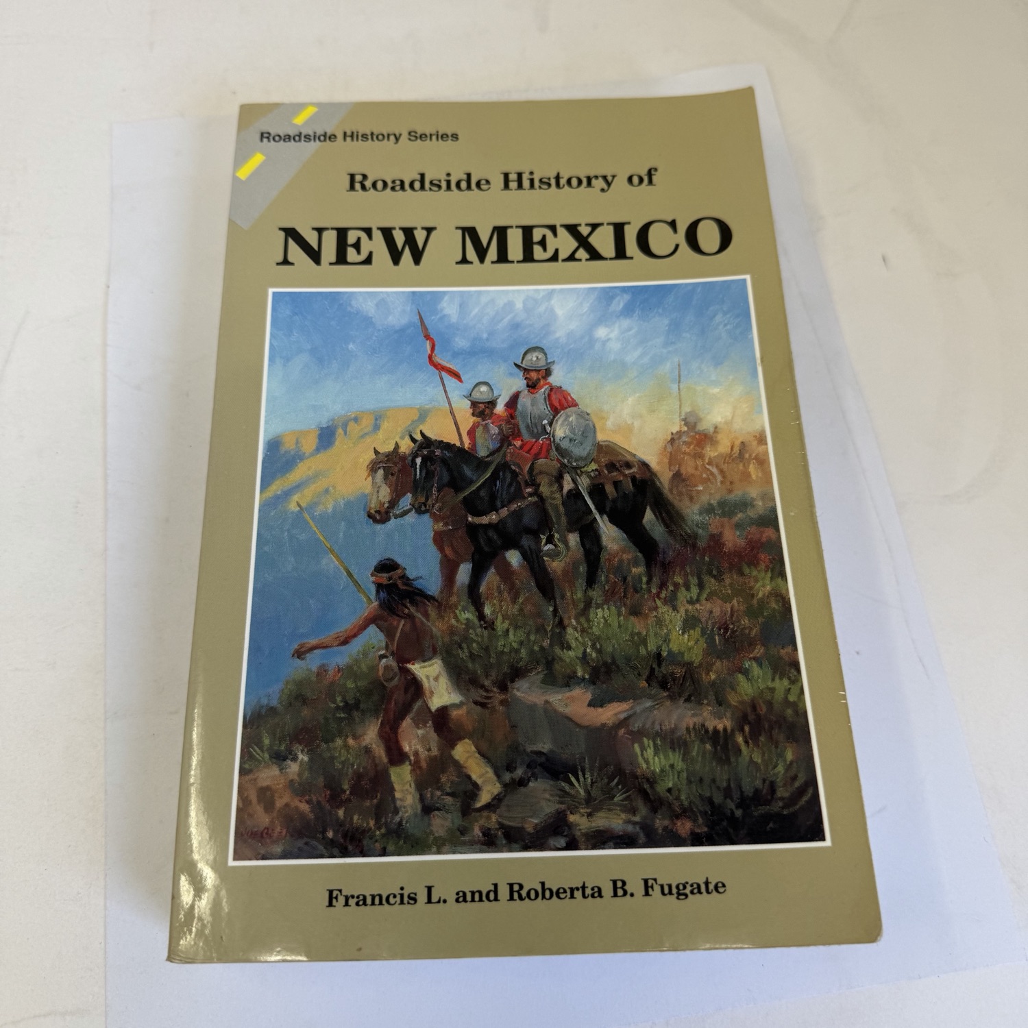 Front cover of Roadside History of New Mexico by Francis L. and Roberta B. Fugate — Roadside History Series header on tan background. Joe Beeler painting of Spanish conquistadors on horseback encountering a Native American in desert landscape with mesa backdrop. Mountain Press Publishing Company.