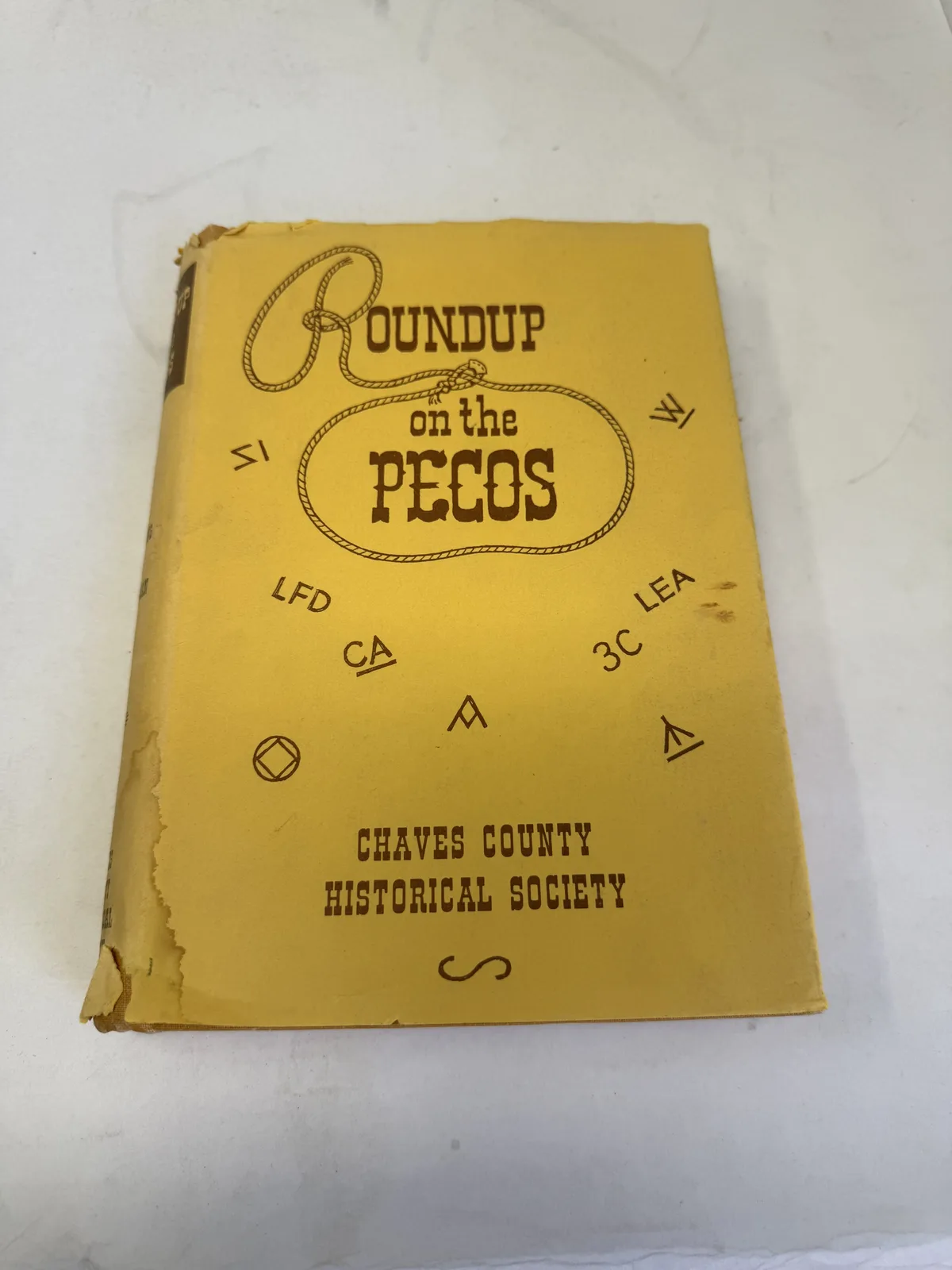 Yellow cover of Roundup on the Pecos, Chaves County Historical Society, lettered in rope script and ringed with historic Pecos Valley cattle brands including LFD, CA, 3C and LEA, photographed at New Mexico Literacy Project