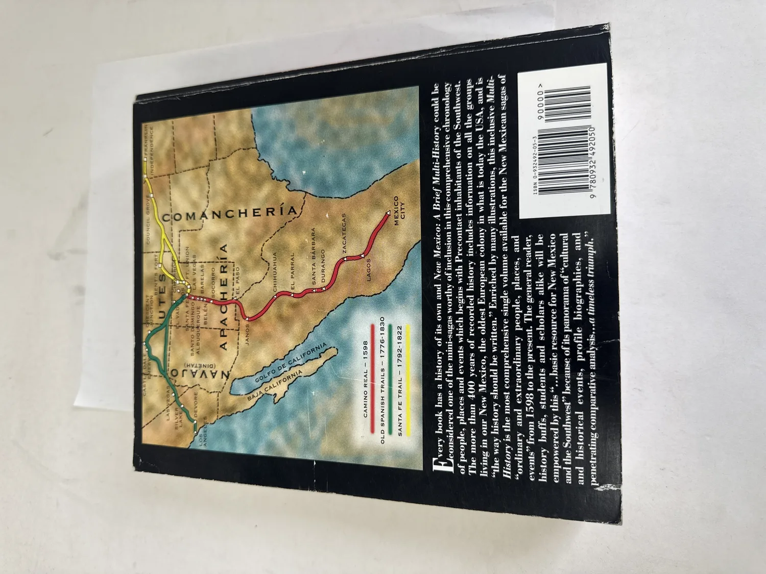 Back cover of New Mexico: A Brief Multi-History showing historical map of colonial New Mexico with Camino Real 1598, Old Spanish Trail 1776-1830, Santa Fe Trail 1792-1822, territorial boundaries for Comanchería, Apachería, Navajo, and Utes, NM towns including Albuquerque, Santa Fe, Socorro, Belén, ISBN barcode. Photographed at the New Mexico Literacy Project sorting desk in Albuquerque.
