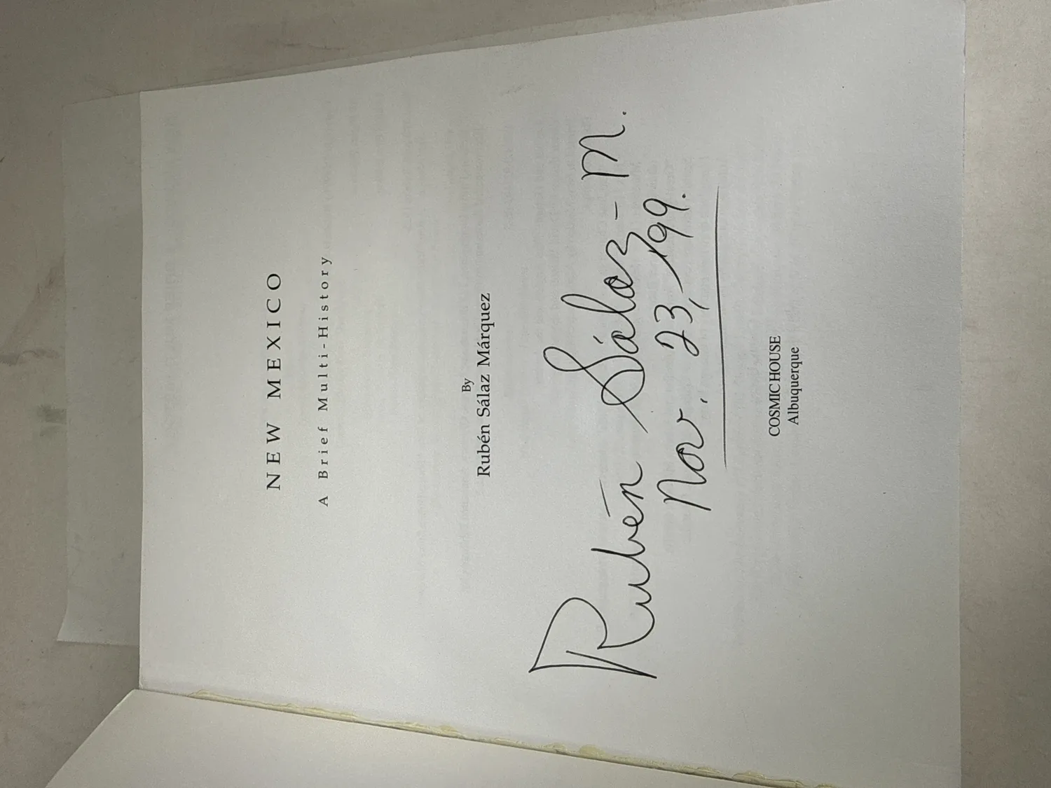 Title page of New Mexico: A Brief Multi-History signed by Rubén Sálaz M. with date Nov. 23, 99 in large confident ink, Cosmic House Albuquerque imprint visible below the signature. Photographed at the New Mexico Literacy Project sorting desk in Albuquerque.