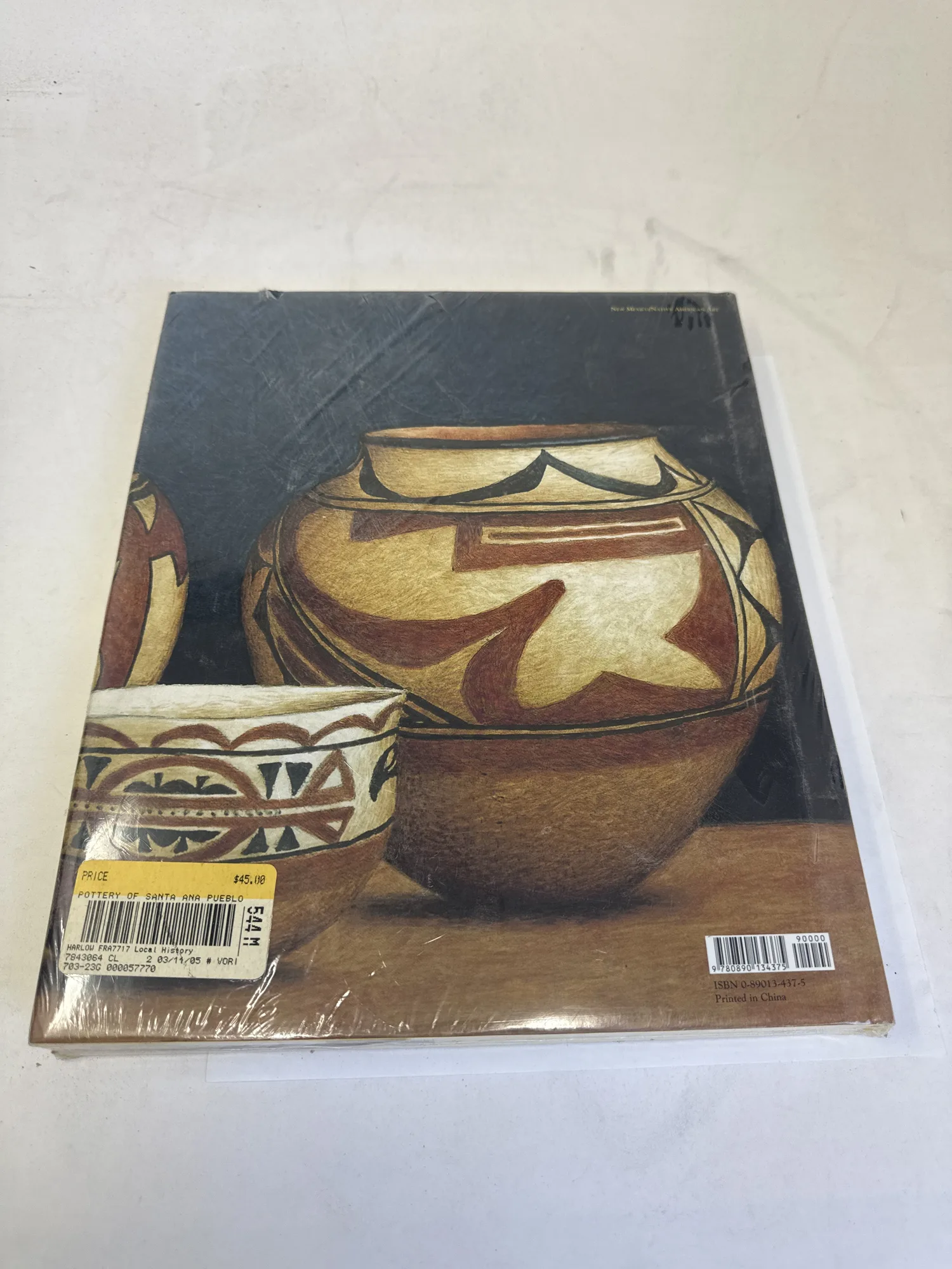 Back cover of The Pottery of Santa Ana Pueblo showing additional polychrome pottery photographs, barcode, price sticker reading forty-five dollars, and library classification tag, still sealed in original shrink wrap. Photographed at the New Mexico Literacy Project sorting desk in Albuquerque.