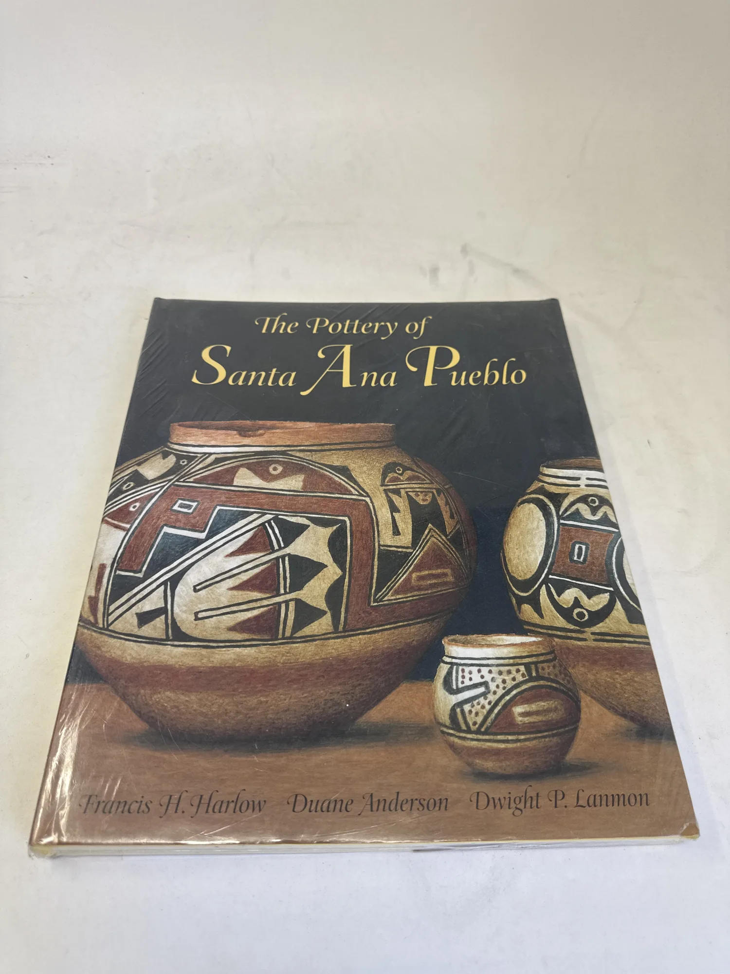 Front cover of The Pottery of Santa Ana Pueblo by Francis H. Harlow, Duane Anderson, and Dwight P. Lanmon, showing polychrome pottery vessels with geometric designs in red, black, and cream, still factory-sealed in original shrink wrap. Photographed at the New Mexico Literacy Project sorting desk in Albuquerque.