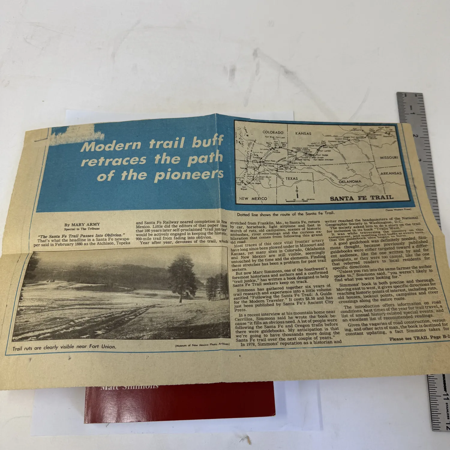 Vintage newspaper clipping found inside a copy of Following the Santa Fe Trail by Marc Simmons — headline reads Modern trail buff retraces the path of the pioneers by Mary Army, with a Santa Fe Trail route map showing the path from Franklin Missouri to Santa Fe New Mexico through Kansas Colorado Oklahoma and Texas, and a photograph of trail ruts near Fort Union New Mexico. Provenance ephemera from an Albuquerque estate donation to the New Mexico Literacy Project.