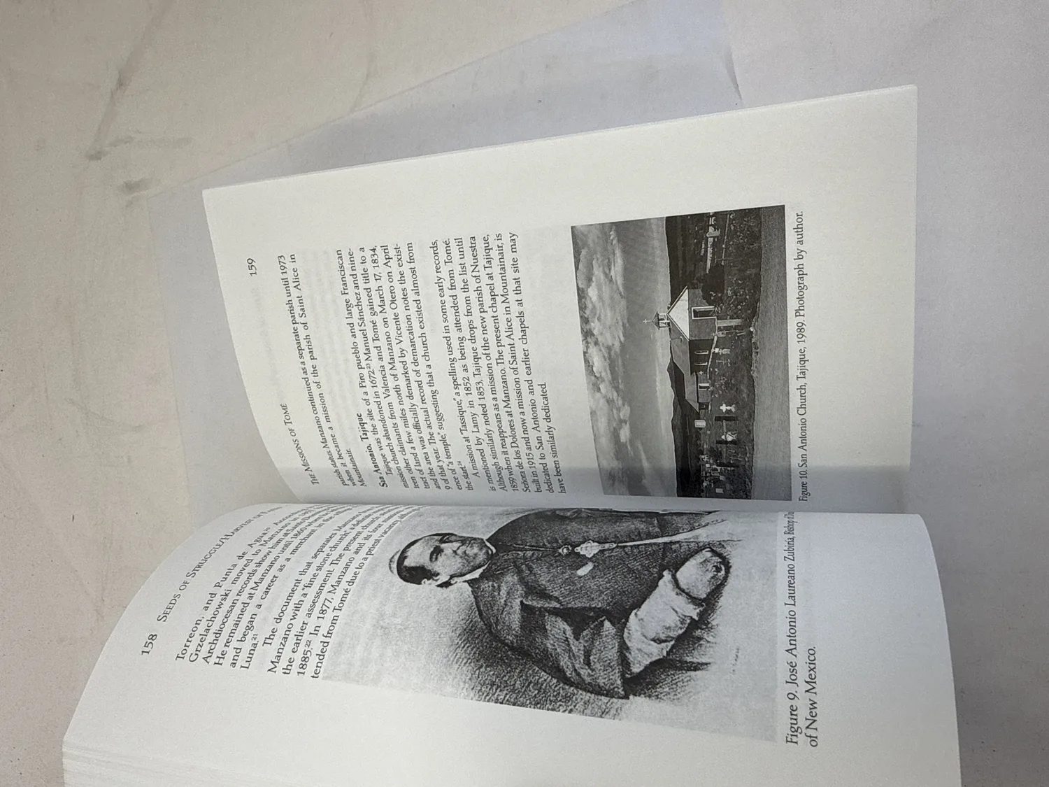 Interior spread of Seeds of Struggle, Harvest of Faith showing pages 158-159 with mission history text on San Antonio de Tajique, portrait of Jos&eacute; Antonio Laureano Zubia, and photograph of San Antonio Church at Tajique 1989. Photographed at the New Mexico Literacy Project sorting desk in Albuquerque.