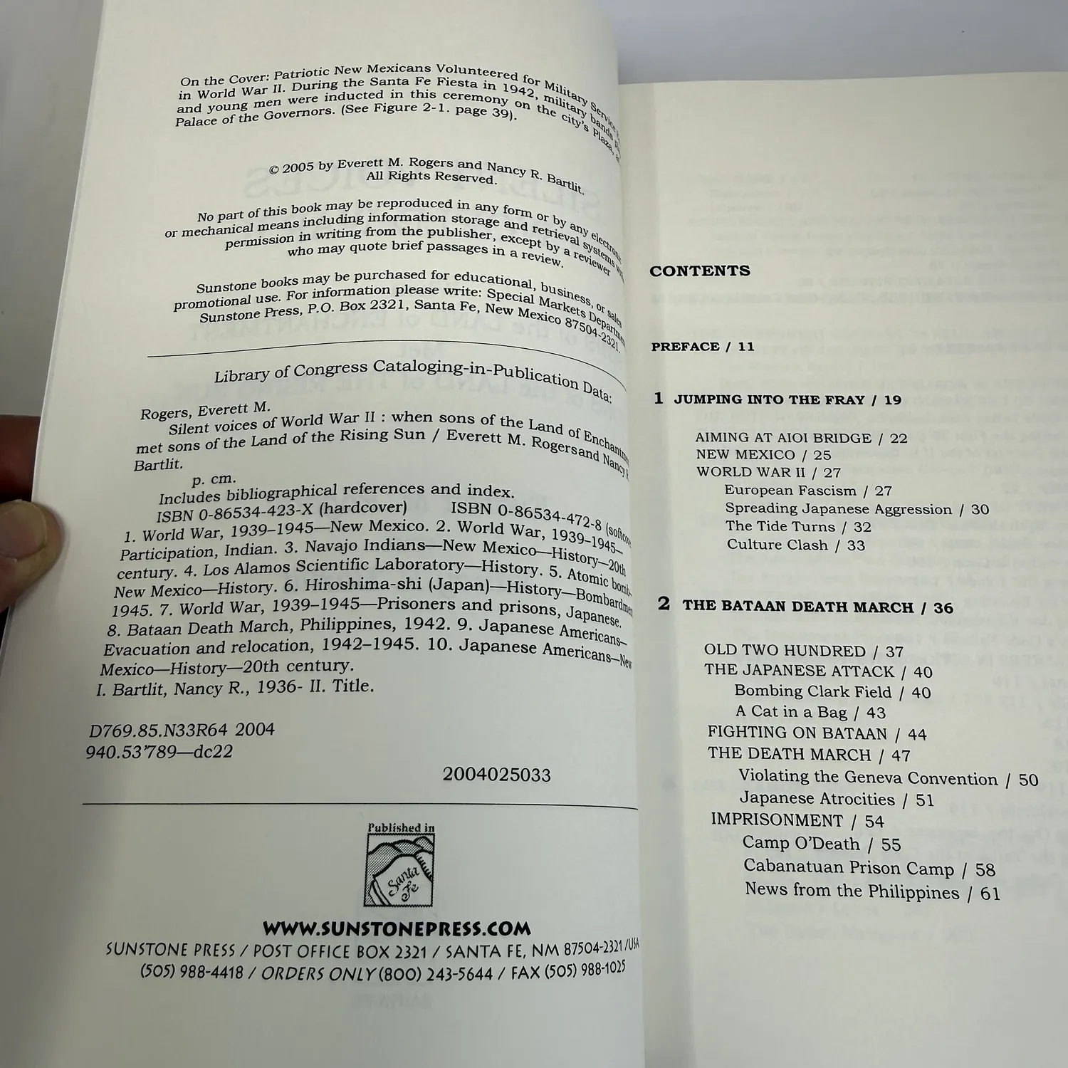 Copyright page and table of contents of Silent Voices of World War II by Rogers and Bartlit showing Sunstone Press Santa Fe imprint, ISBN 0-86534-423-X hardcover, ISBN 0-86534-472-8, chapter listings covering Bataan Death March 200th Coast Artillery Los Alamos Manhattan Project Navajo code talkers Hiroshima bombing and Japanese American internment camp near Santa Fe, photographed at New Mexico Literacy Project sorting desk