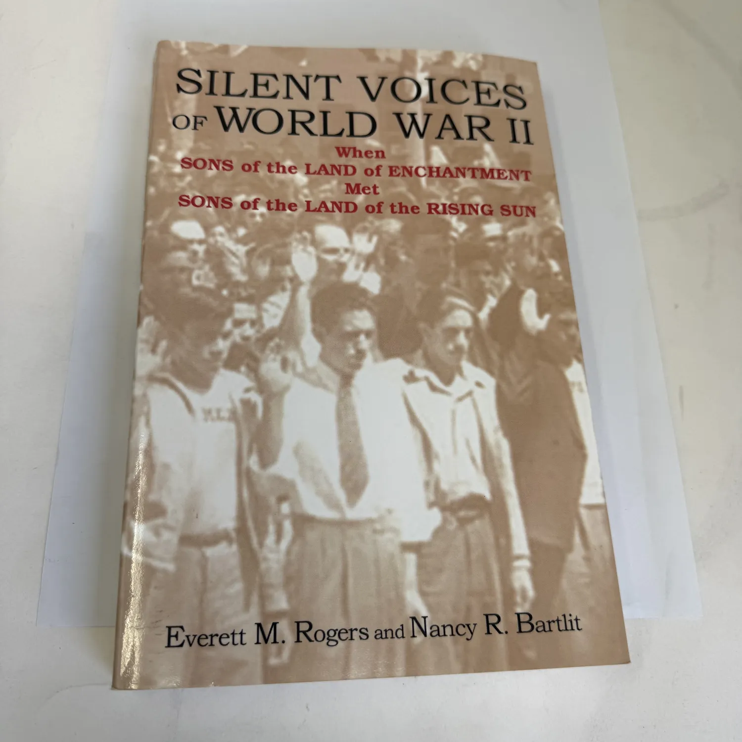 Front cover of Silent Voices of World War II When Sons of the Land of Enchantment Met Sons of the Land of the Rising Sun by Everett M. Rogers and Nancy R. Bartlit, Sunstone Press Santa Fe 2005, hardcover with composite image of New Mexico WWII scenes including Bataan Death March soldiers and Los Alamos Manhattan Project landscape, photographed at the New Mexico Literacy Project sorting desk in Albuquerque