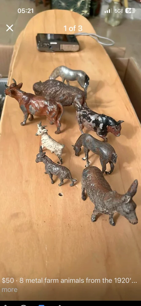 A phone screenshot of a Facebook Marketplace listing showing eight small metal farm animal figurines from the 1920s — horse, cow, pig, goat, sheep, dog, all in cast metal — listed at fifty dollars, photographed on a wooden surface alongside what appears to be a vintage camera.