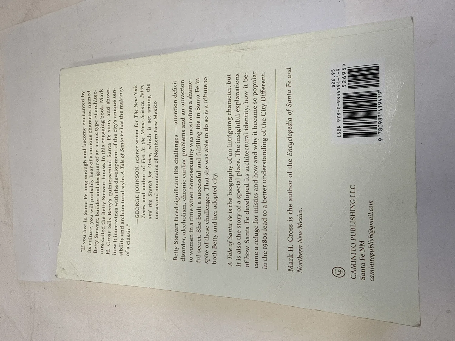 Back cover of A Tale of Santa Fe by Mark H. Cross showing George Johnson blurb, book description mentioning LGBT themes, ISBN 978-0-9834194-1-9, Caminito Publishing LLC Santa Fe NM. Photographed at the New Mexico Literacy Project sorting desk in Albuquerque.