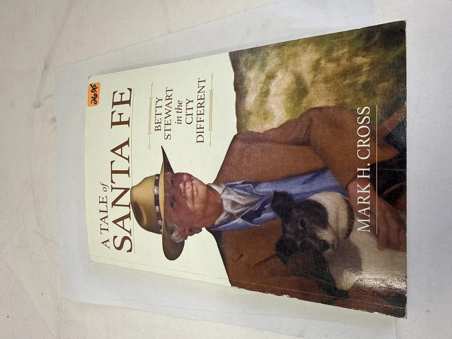 Front cover of A Tale of Santa Fe: Betty Stewart in the City Different by Mark H. Cross — oil portrait by George Mandus showing Betty Stewart in cowboy hat with dog, southwestern palette, orange thrift-store price sticker visible. Photographed at the New Mexico Literacy Project sorting desk in Albuquerque.