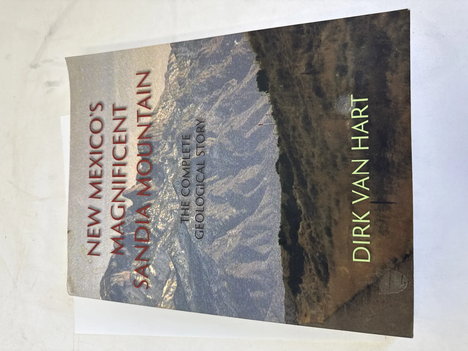 Front cover of New Mexico's Magnificent Sandia Mountain: The Complete Geological Story by Dirk Van Hart &mdash; dramatic Sandia Mountain landscape photograph from US 566 showing rugged ridgeline and geological formations, Sunstone Press 2023. Photographed at the New Mexico Literacy Project sorting desk in Albuquerque.