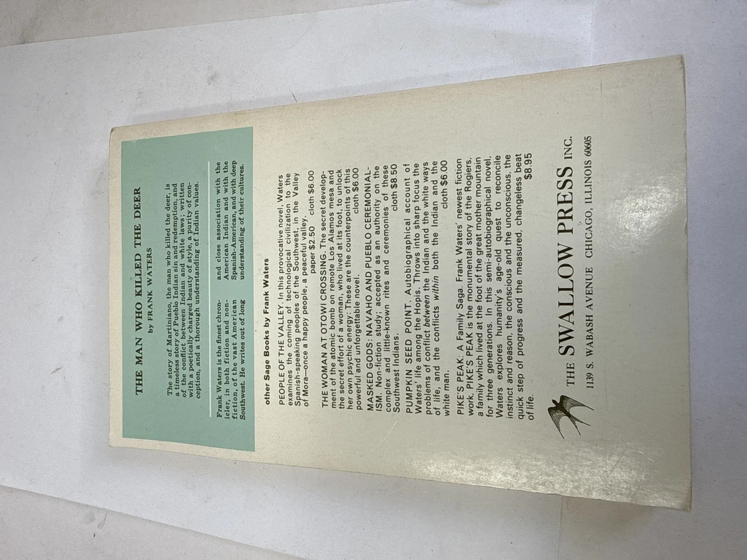 Back cover of The Man Who Killed the Deer Sage Paperbook edition listing other Frank Waters titles including People of the Valley, The Woman at Otowi Crossing, Masked Gods, Pumpkin Seed Point, and Pike's Peak with the Swallow Press swallow-bird colophon and 1139 S. Wabash Avenue Chicago address. Photographed at the New Mexico Literacy Project sorting desk in Albuquerque.