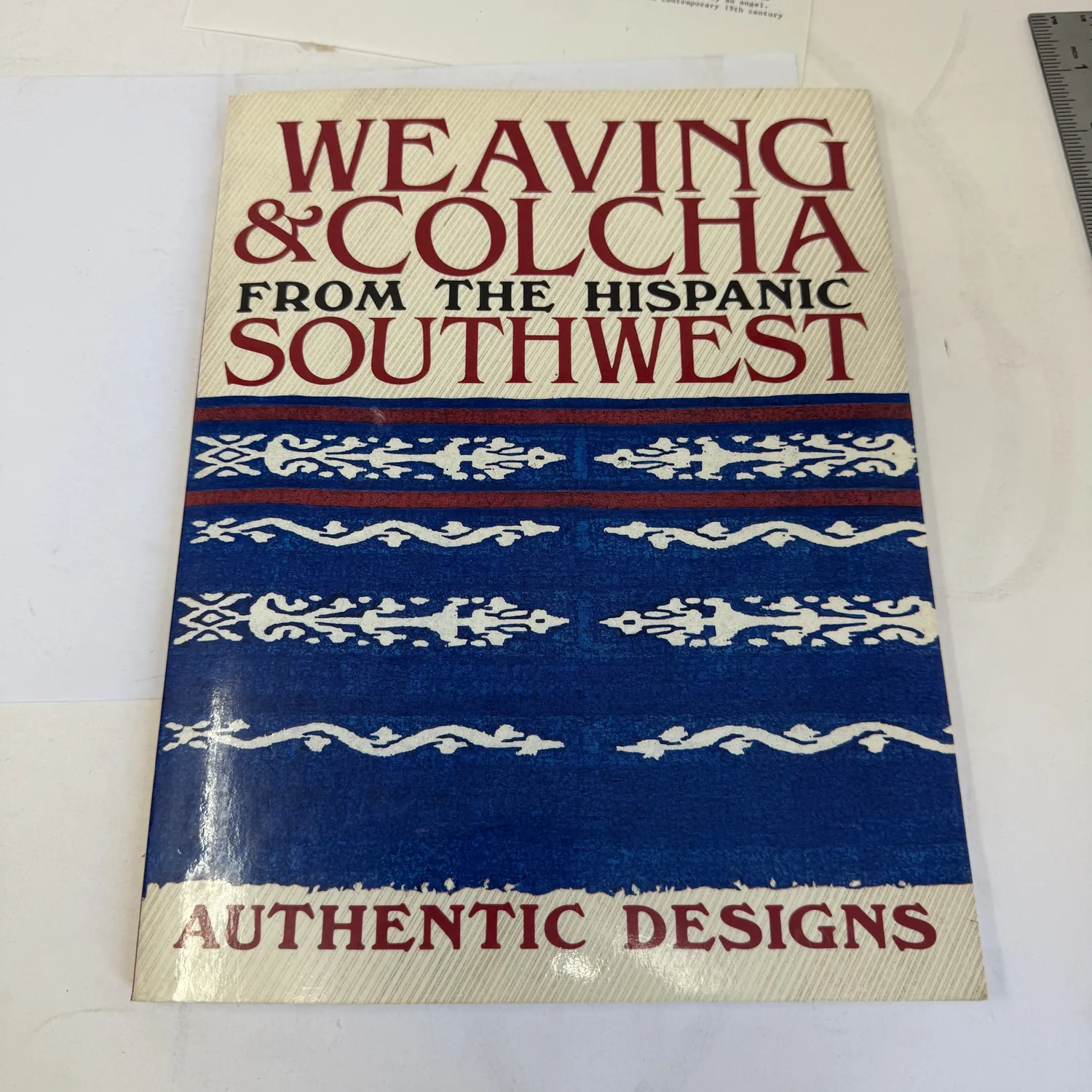 First edition cover of Weaving and Colcha from the Hispanic Southwest Authentic Designs edited by William Wroth, Ancient City Press Santa Fe 1985, showing blue and red Hispanic weaving design pattern on cream background, photographed at New Mexico Literacy Project