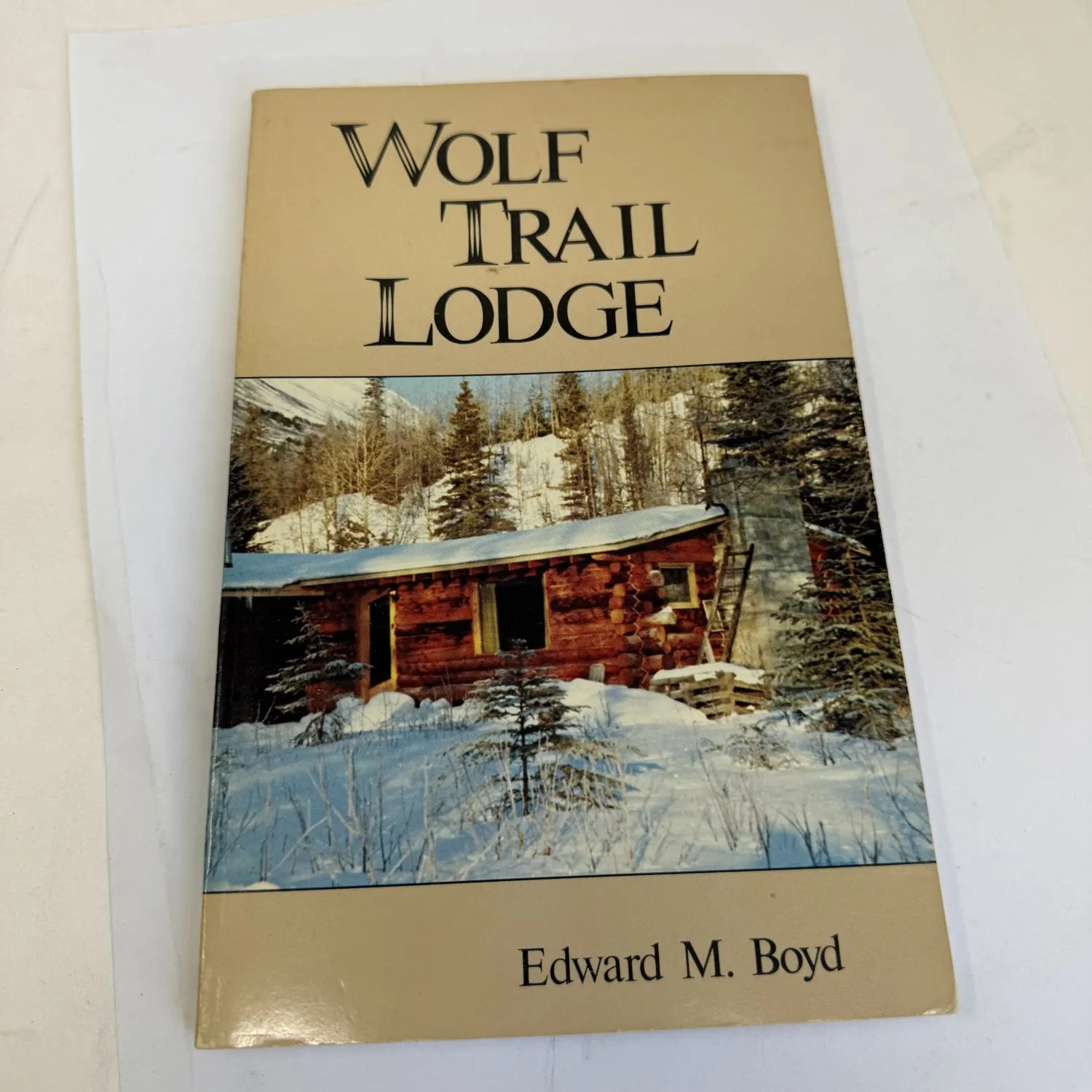 Front cover of Wolf Trail Lodge by Edward M. Boyd, Alaska Northwest Publishing Company 1984, showing a snow-covered log cabin in the Kenai Peninsula spruce forest with mountains behind, tan cover with black title lettering. Found in a New Mexico estate cleanout by the New Mexico Literacy Project.