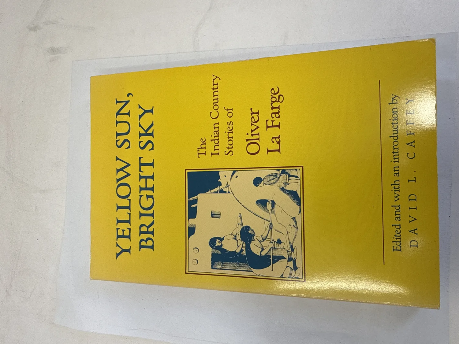 Front cover of Yellow Sun, Bright Sky: The Indian Country Stories of Oliver La Farge — bright yellow softcover with blue text, line drawing of a Native American scene, edited by David L. Caffey, University of New Mexico Press 1988 First Edition. Photographed at the New Mexico Literacy Project sorting desk in Albuquerque.