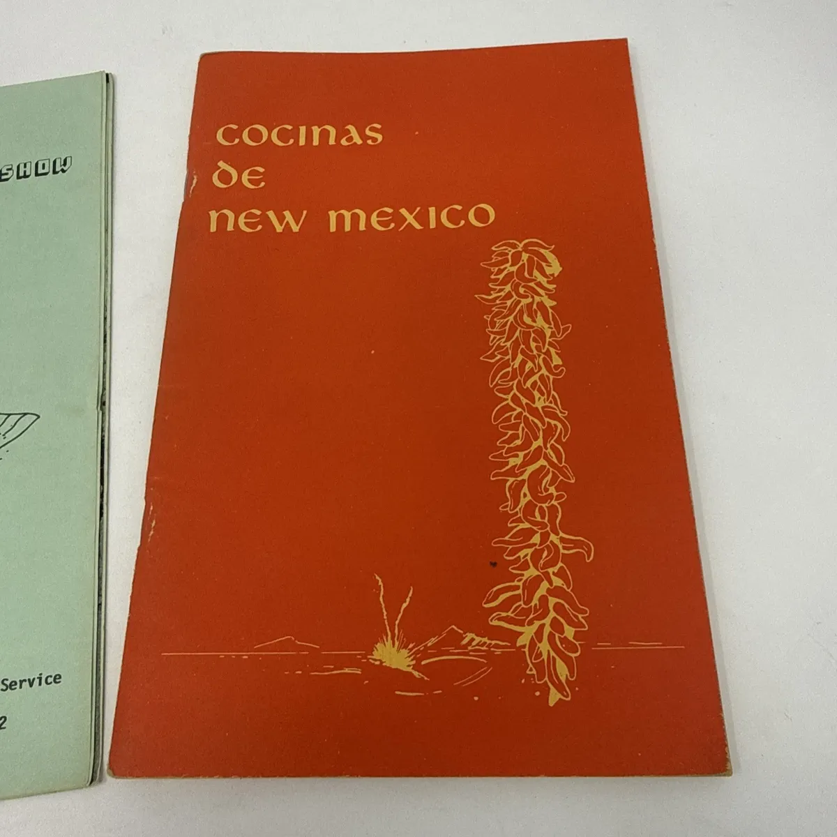 Orange and green Cocinas de New Mexico cookbooks lying overlapping, the orange one with a chile ristra illustration, the green one a Bernalillo County Extension Service publication.