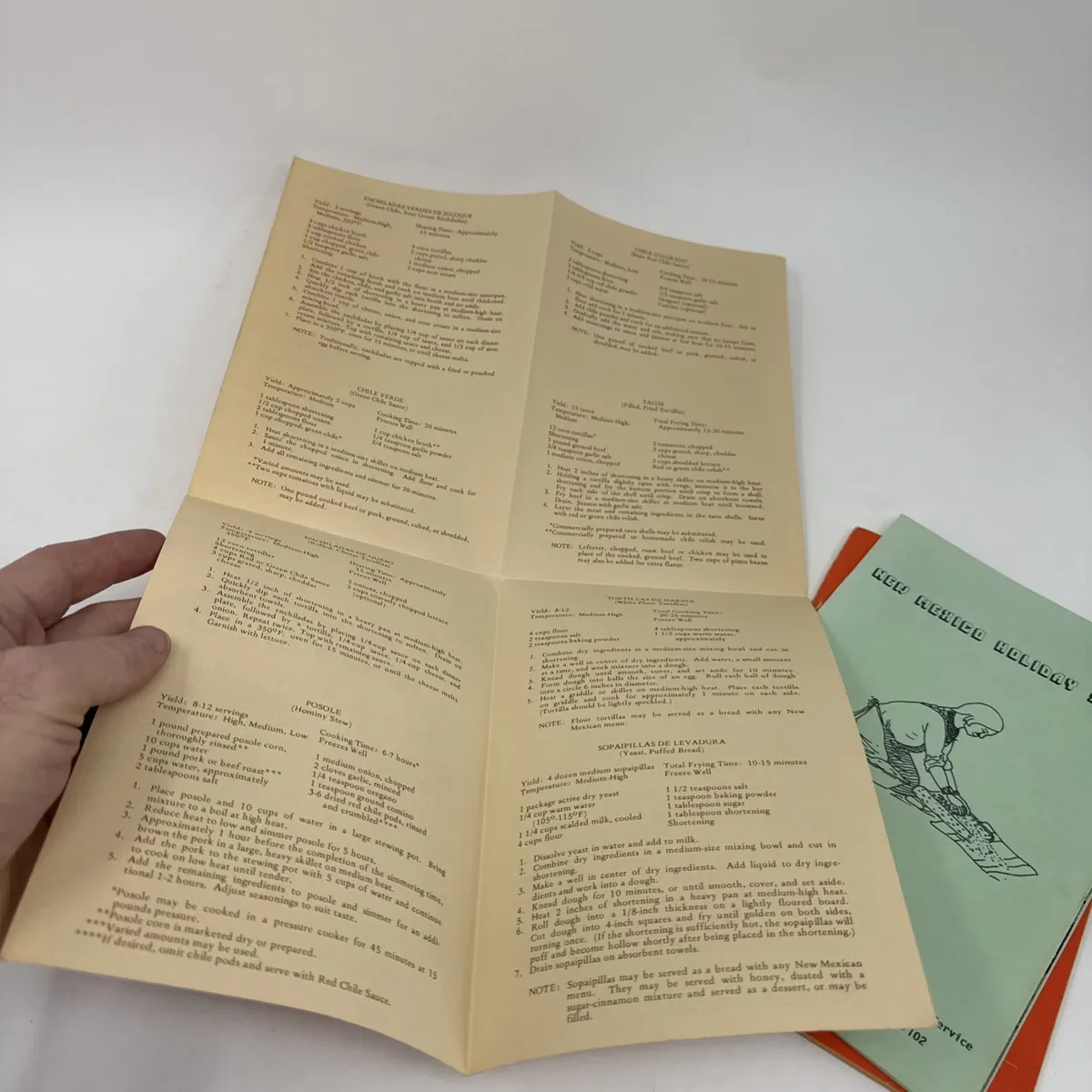 The interior of one of the Cocinas de New Mexico cookbooks held open by hand, showing recipes for posole, chimichangas, capirotada, sopaipillas, and other traditional New Mexican dishes printed in mid-century typewriter typeface on cream-colored paper, with the orange chile-ristra cover of another Cocinas variant visible at the right edge of the frame.