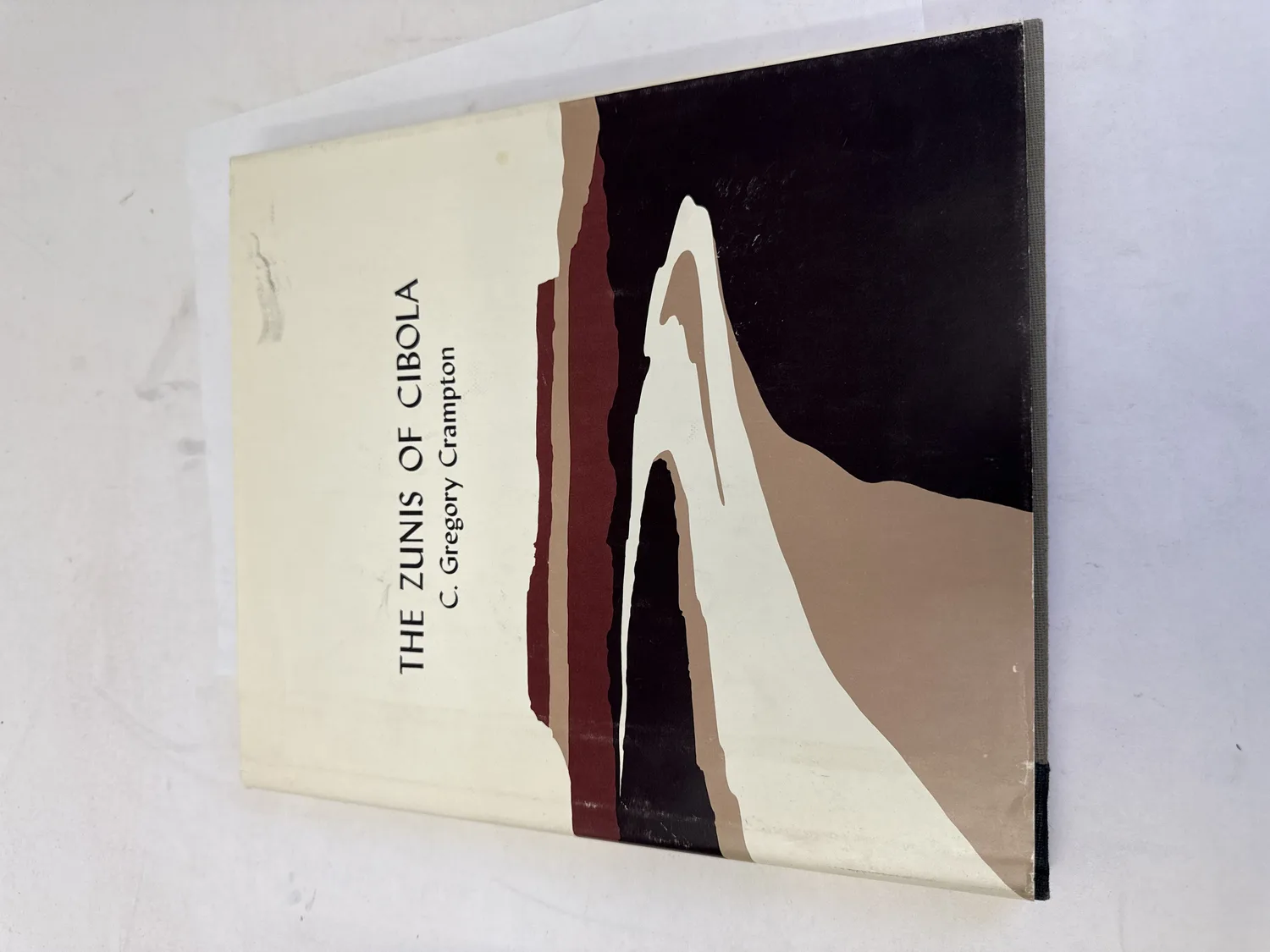 Front cover and dust jacket of The Zunis of Cibola by C. Gregory Crampton, 1977 first edition, featuring an abstract landscape design in black, tan, burgundy, and cream evocative of western New Mexico mesa and canyon country. Photographed at the New Mexico Literacy Project sorting desk in Albuquerque.